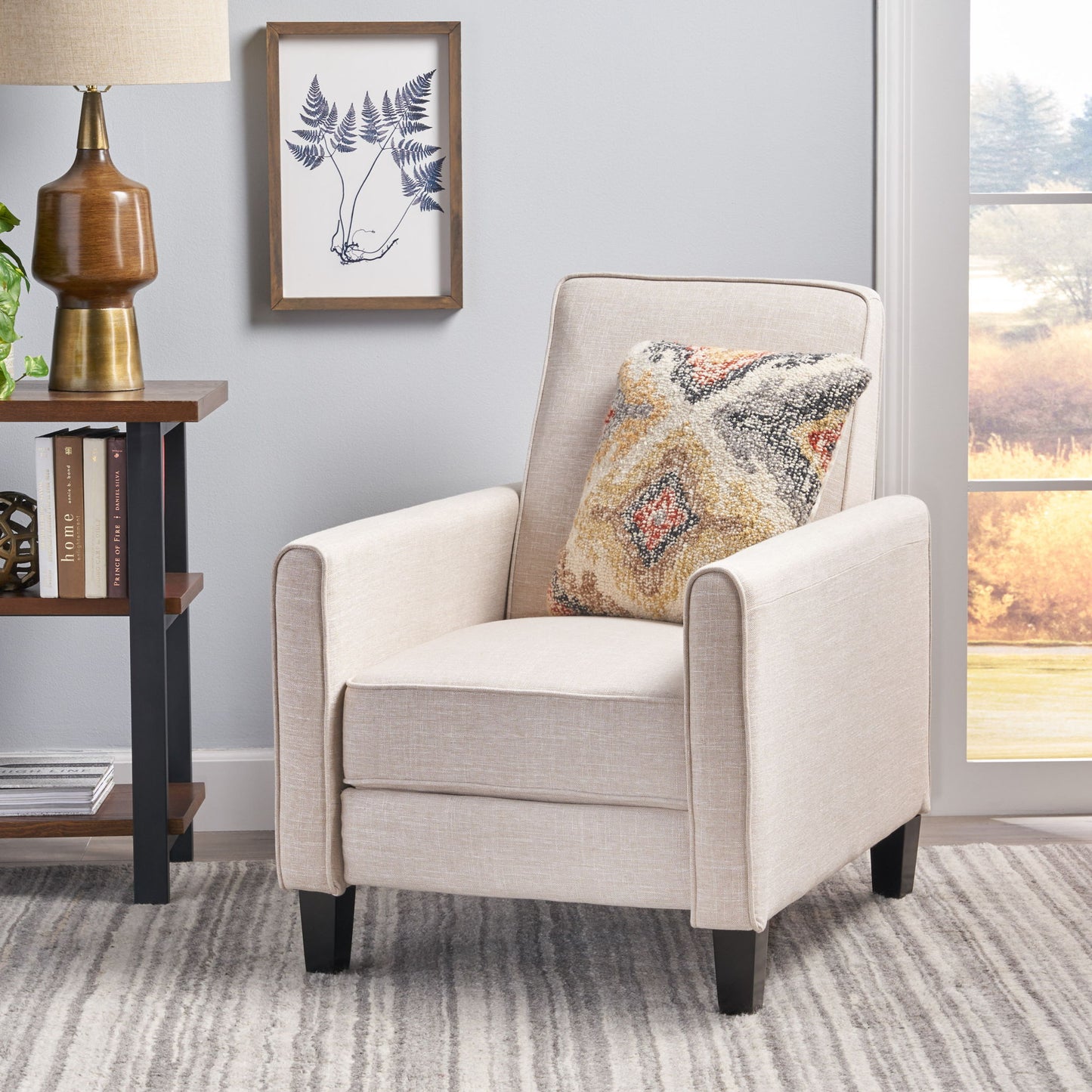 Beige armchair with a patterned pillow in a room with a lamp, books, and a framed picture on the wall.