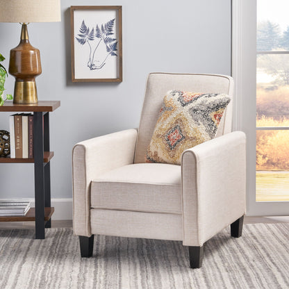 Beige armchair with a patterned pillow in a room with a lamp, books, and a framed picture on the wall.