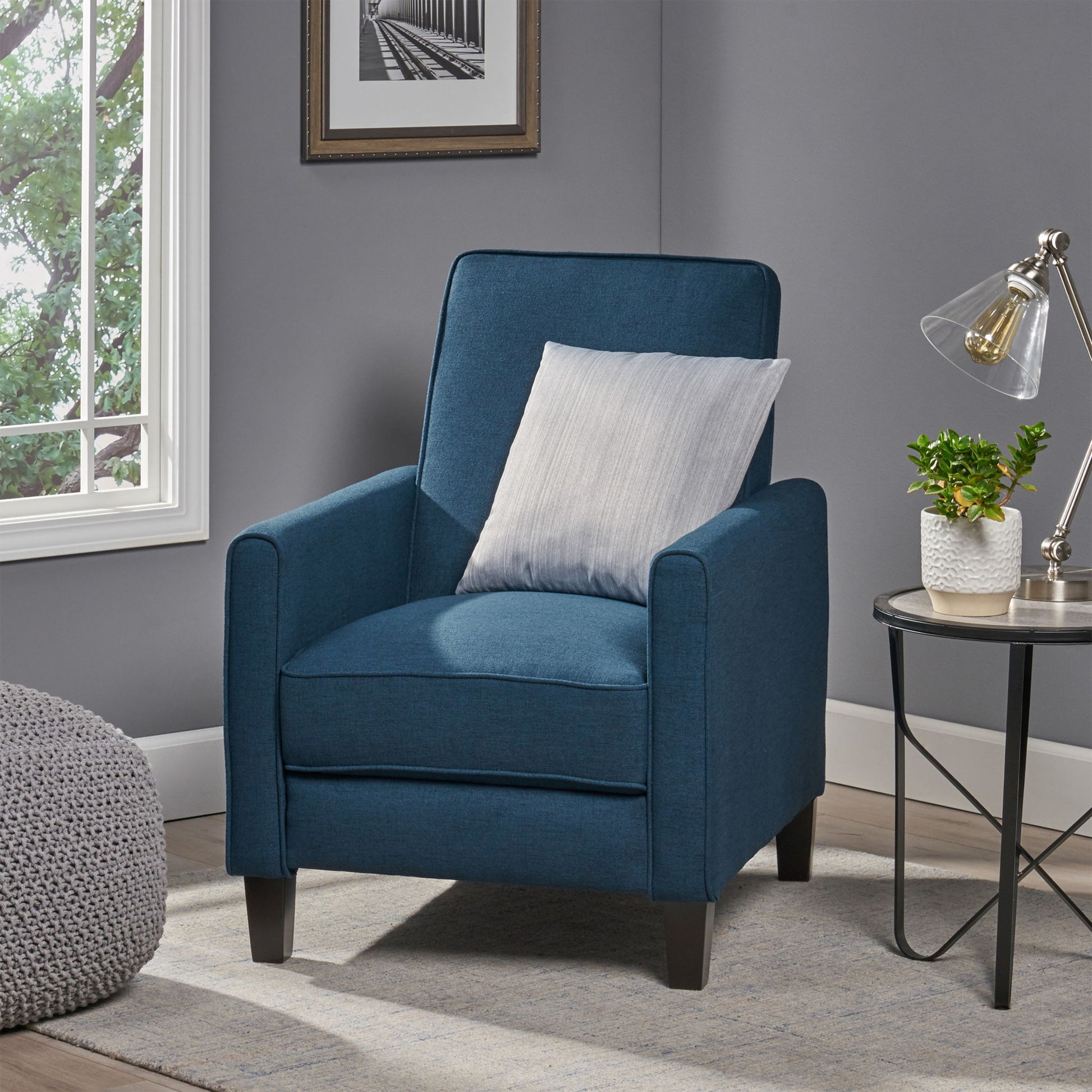 Blue armchair with a gray pillow in a room with a window and side table.